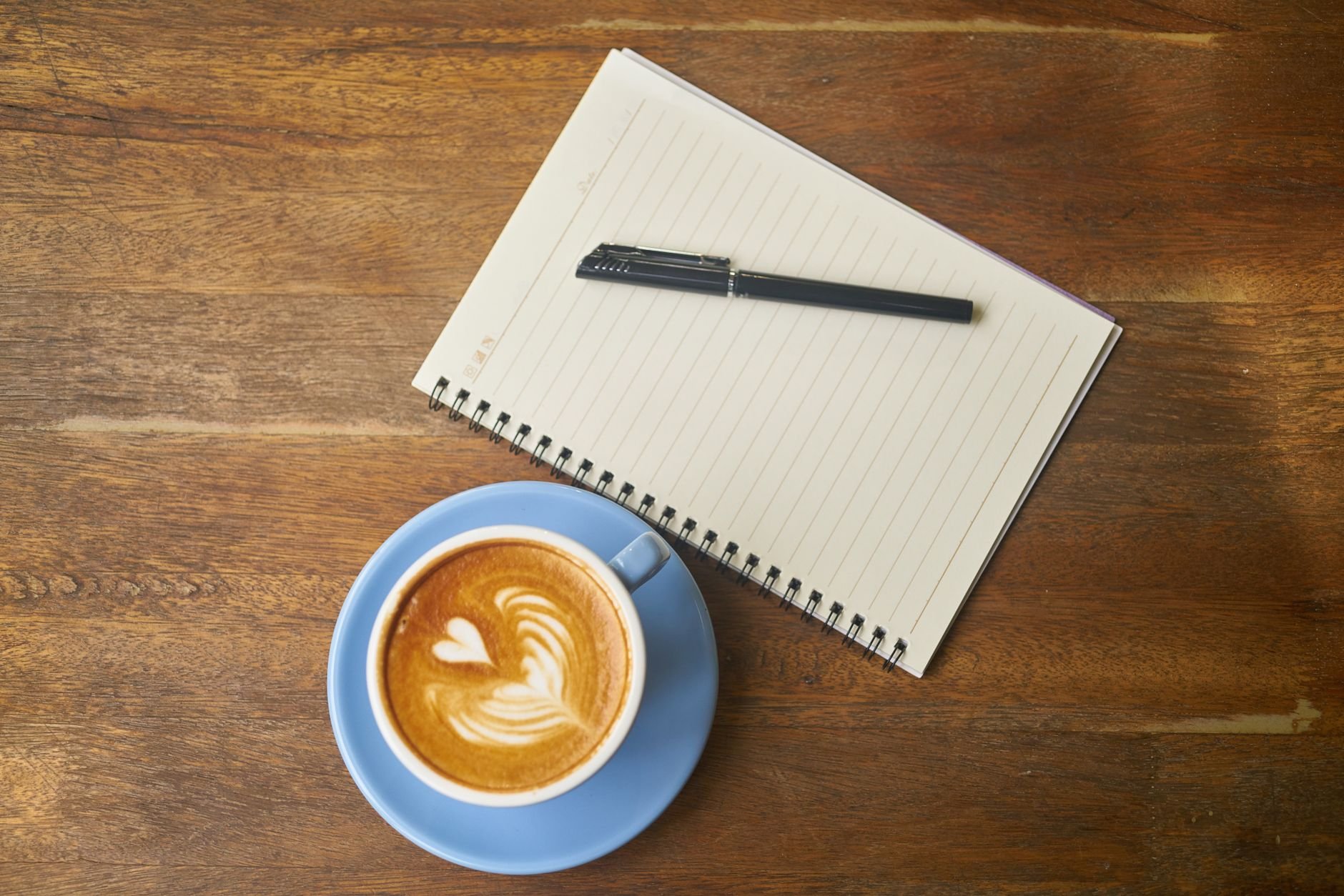 Aerial view of a coffee cup with latte art beside a notebook and pen on a wooden desk.